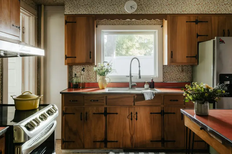 Cuisine en bois foncé sous une fenêtre : placards à cadre avec ferrures noires, évier inox, crédence à motifs et bouquet de fleurs.