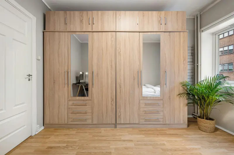 Grande armoire en chêne clair occupant tout un mur, avec deux portes miroirs, des tiroirs en bas et une plante près de la fenêtre.