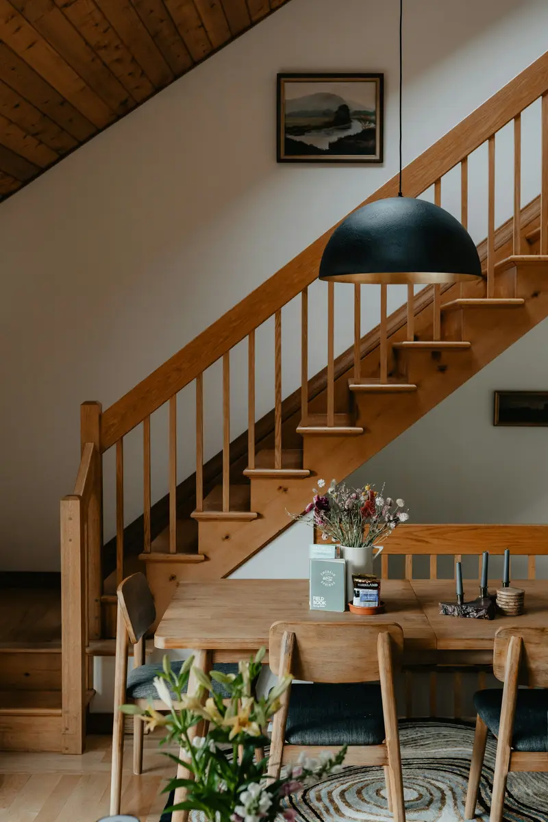 Escalier droit en bois blond avec rampe à balustres, vu depuis une salle à manger où une suspension noire éclaire un bouquet.